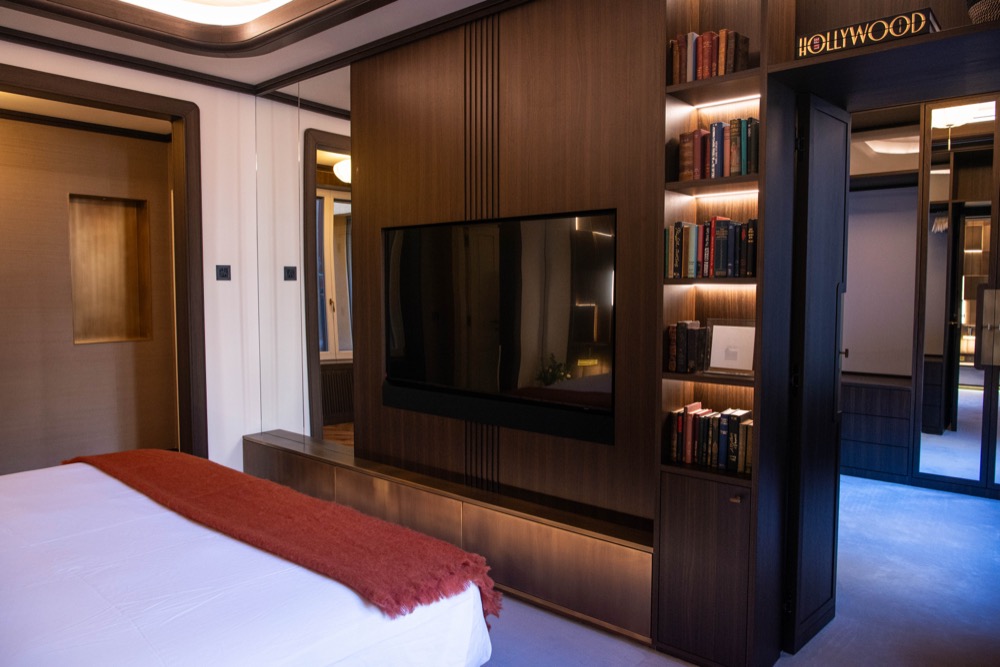 Master bedroom with a white bed, a rust-red throw, a television, bookshelves and a dressing room beyond