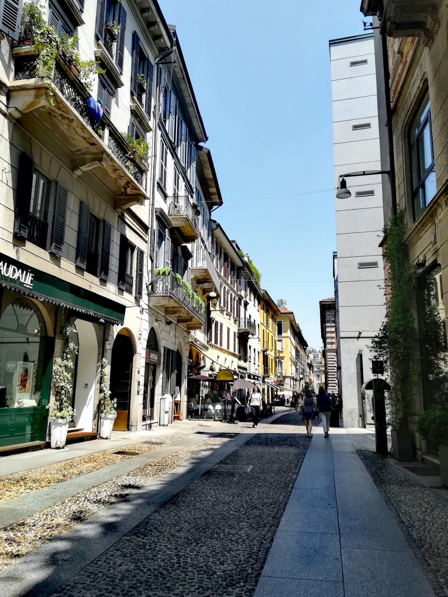 Cobbled Via Fiori Chiari in daylight, with cafés, shuttered windows and planted balconies